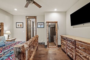 Un dormitorio con cama, cabecera de madera, mesita de noche, lámpara y televisor montado en la pared.