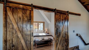 Un dormitorio con un amplio zaguán de madera corredera, una cama con ropa blanca, un banco y una ventana con hojas de otoño visibles desde el exterior.
