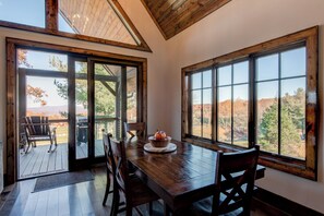 A dining area with a wooden table and chairs, large windows with a view of trees and hills, and a wooden deck outside. - Northern Exposure at Eagles Nest (Elk Park)