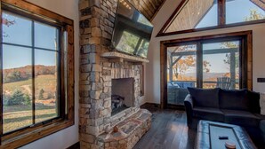 Una chimenea de piedra con televisión, un sofá de cuero y amplios ventanales con vistas al campo.