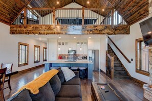 Amplio espacio tipo loft con techo alto, vigas de madera a la vista, cocina central con armarios blancos y chimenea de piedra.