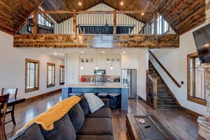 Amplio espacio tipo loft con techo alto, vigas de madera a la vista, cocina central con armarios blancos y chimenea de piedra.