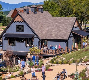 A large, rustic building with a sloped roof and multiple windows. There is a deck with people sitting and standing, surrounded by landscaped gardens and rocks. - Wine Down at Eagles Nest (Elk Park)