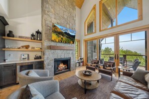 A spacious living room with a stone fireplace, comfortable seating, a kitchen area with modern appliances, and large windows offering a view of the outdoors. - Wine Down at Eagles Nest (Elk Park)