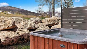 A hot tub with a wooden exterior and a stone top, surrounded by large rocks and a wooden fence, with a mountainous landscape in the background. - 6486 Lakeside #15 (Huntsville)