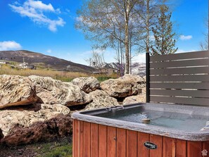 Ett bubbelbad med en träpanel på utsidan och en topp av sten, omgivet av stora stenar och ett trästaket, med ett bergigt landskap i bakgrunden.