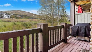 Una terraza de madera con una parrilla y una mesa cubierta, con vistas a un campo de césped y una montaña a lo lejos.