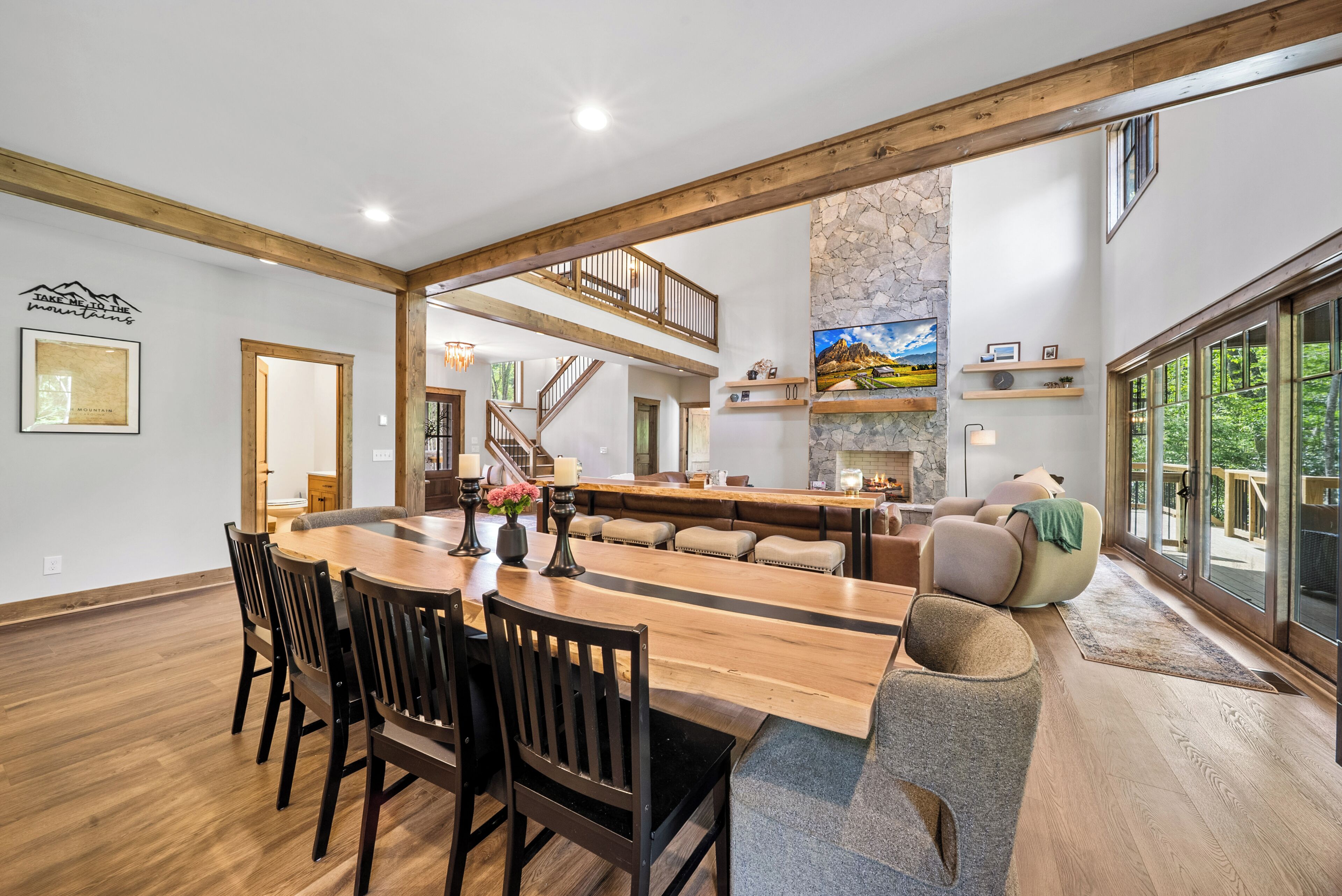 Un comedor amplio con una larga mesa de madera y sillas, una cocina con detalles en pared de piedra, y vistas al exterior a través de amplias ventanas.