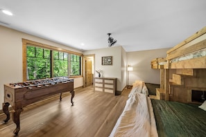Ein Zimmer mit Kicker, einem Bett mit Holz Kopfteil, einem Fenster mit Blick auf Bäume und einem Deckenventilator.