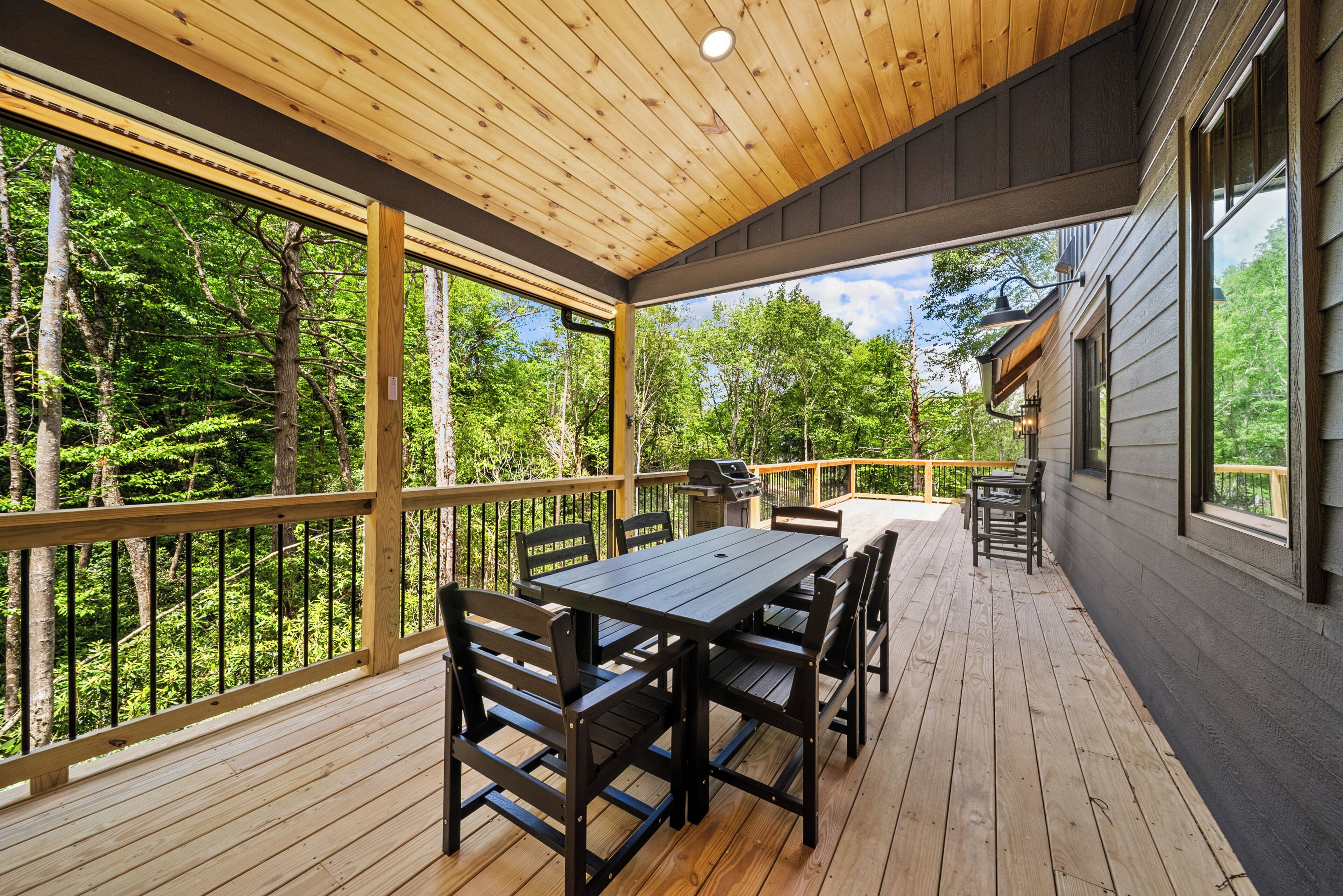 A covered deck with a wooden table and chairs, overlooking a forested area.