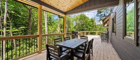 A covered deck with a wooden table and chairs, overlooking a forested area.