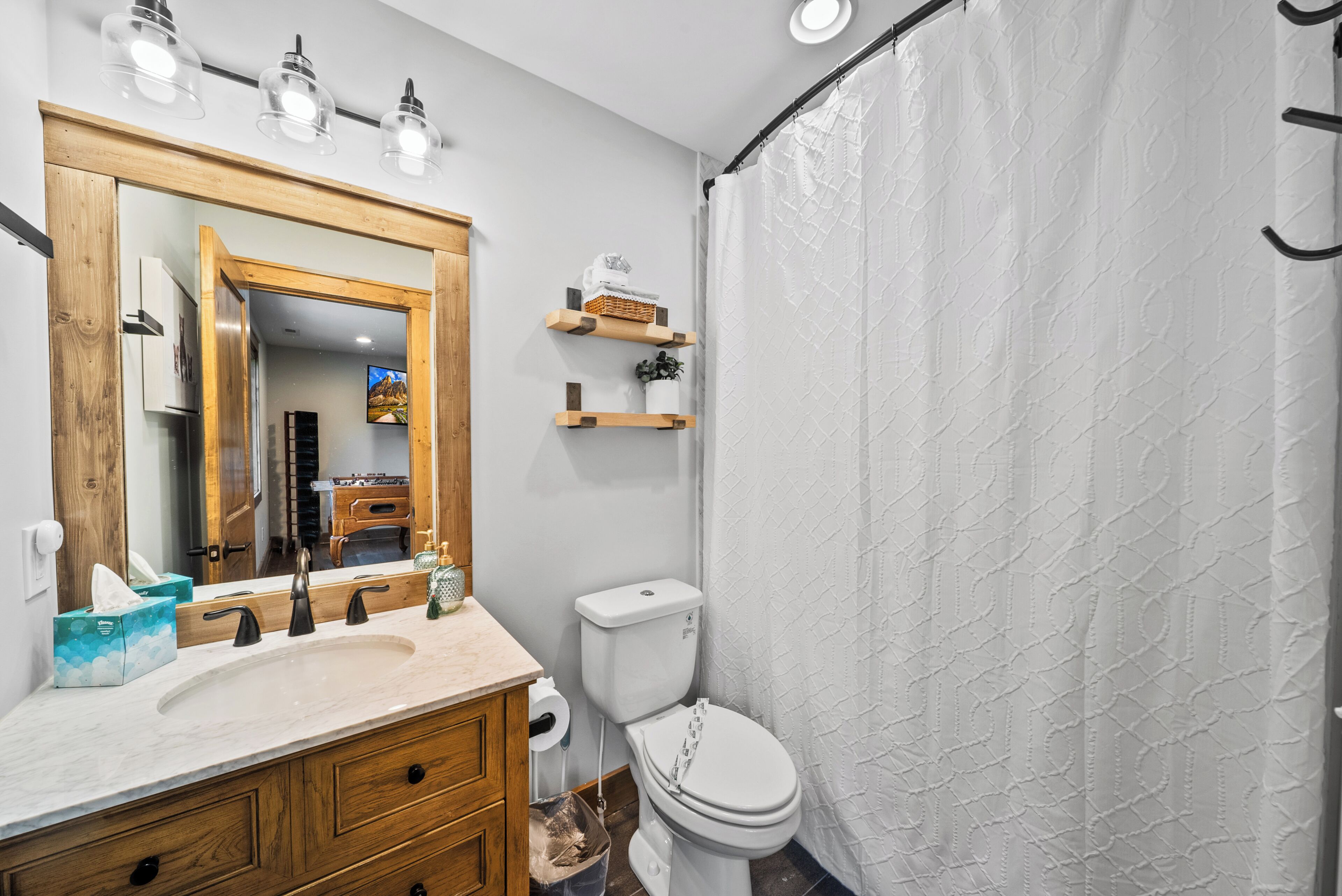 Une salle de bain avec un meuble-lavabo en bois, des toilettes, un rideau de douche et un miroir avec éclairage intégré.