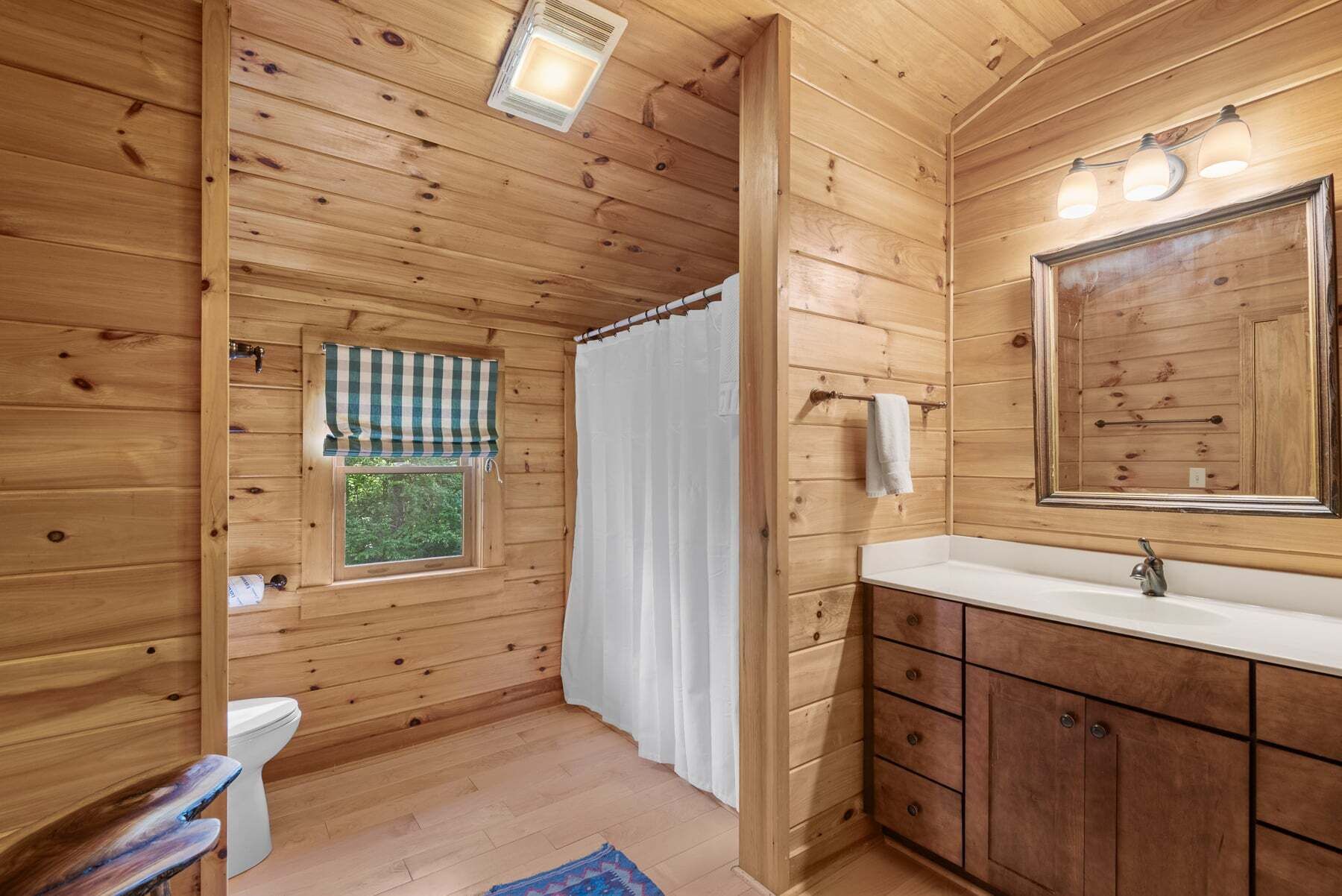 A wooden cabin bathroom with a toilet, sink, mirror, and a window with a checkered curtain.