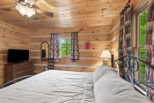 Un dormitorio de cabaña de madera con cama, televisor, cómoda, escalera, ventilador de techo y ventana con cortinas.