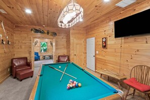 A wooden interior with a pool table, two leather chairs, a television, and a clock on the wall.