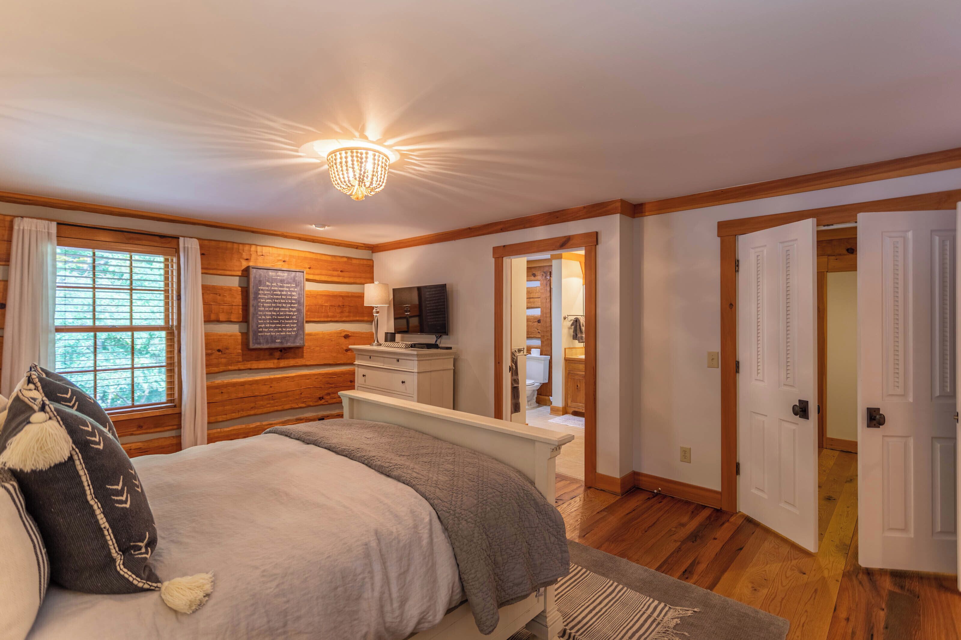 A bedroom with a wooden wall, a bed with a grey blanket, a bedside table, and a chandelier.