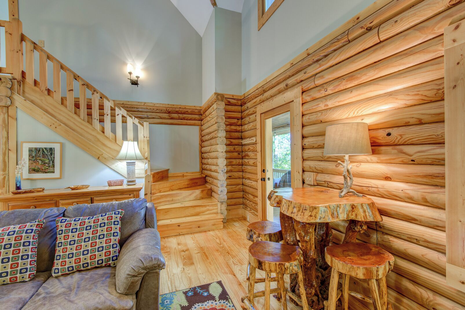 L’intérieur d’une cabane en rondins, avec un escalier, un canapé agrémenté de coussins à motifs, une table avec une lampe et une chaise en bois rustique.