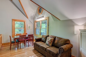 A cozy room with a wooden dining table and chairs, a leather sofa, and large windows offering a view of greenery. - Hideaway Homestead (Banner Elk)