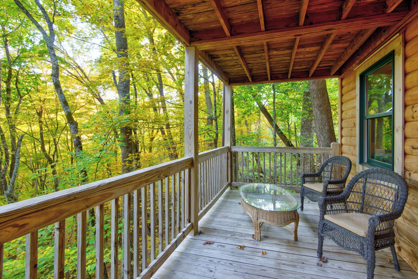 A wooden deck with a glass-top table and wicker chairs, surrounded by a forest.