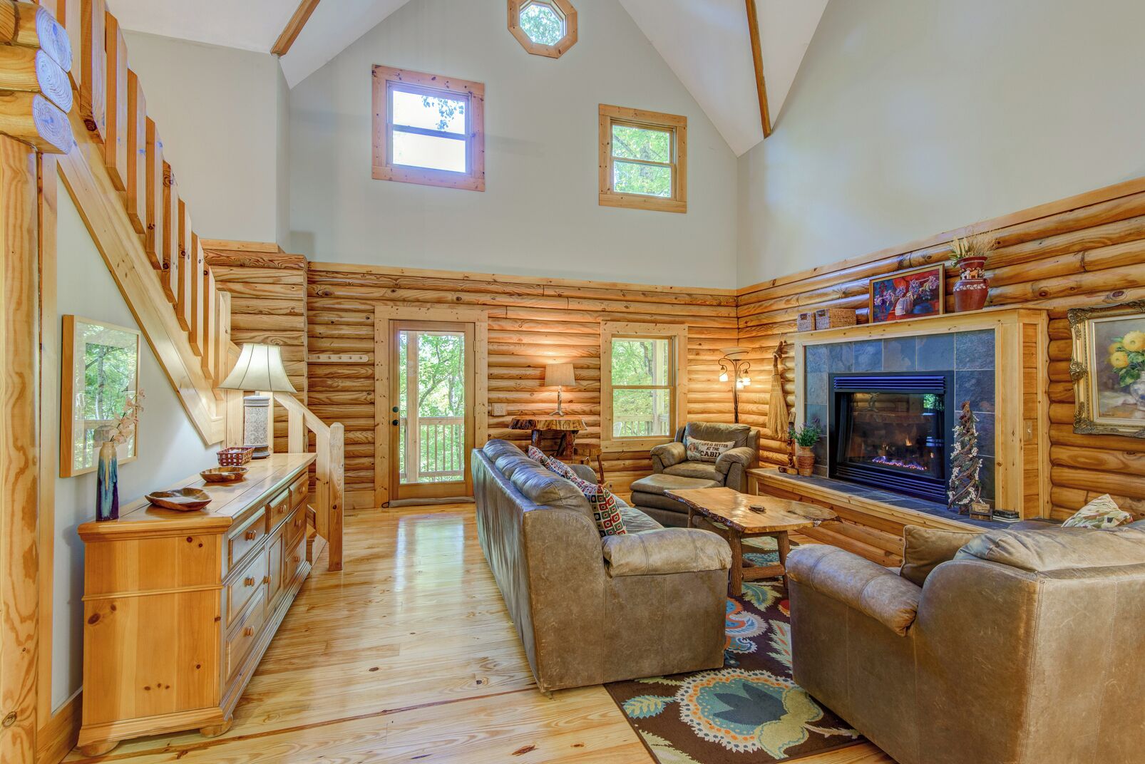A log cabin interior with a wooden staircase, a fireplace, a sofa, and a dining area.
