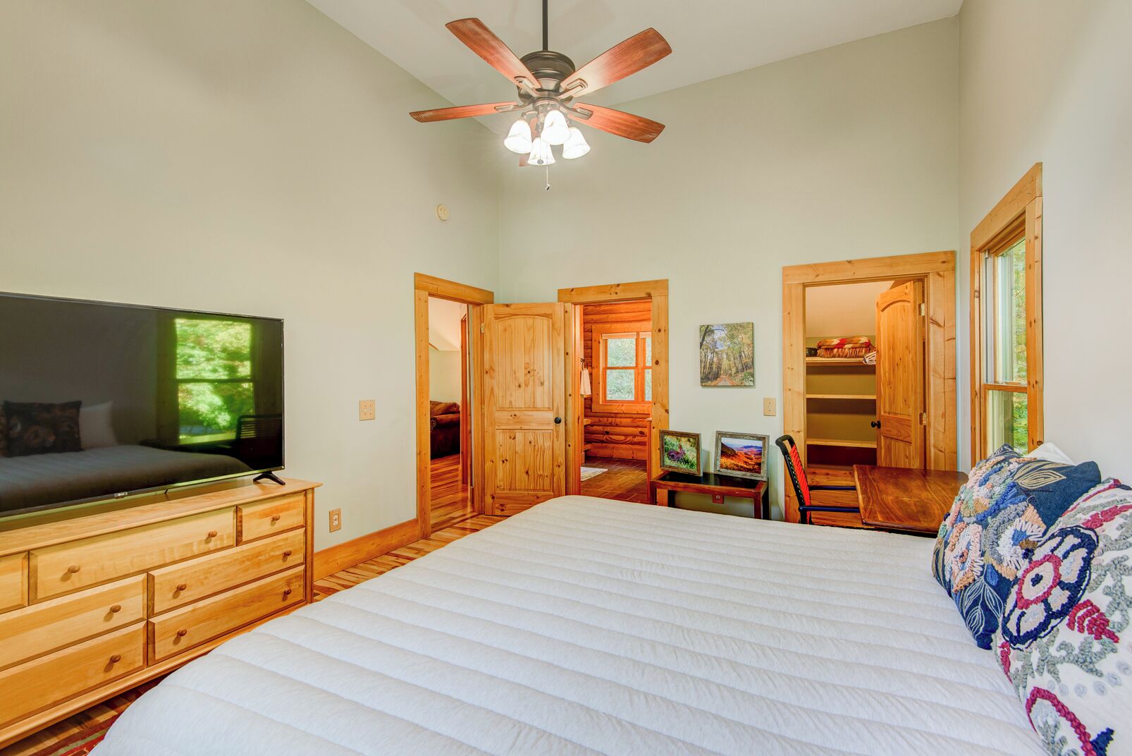 Ein Schlafzimmer mit Bett, einem Holzschrank, Fernseher, Deckenventilator und Blick auf einen Wald durch ein Fenster.