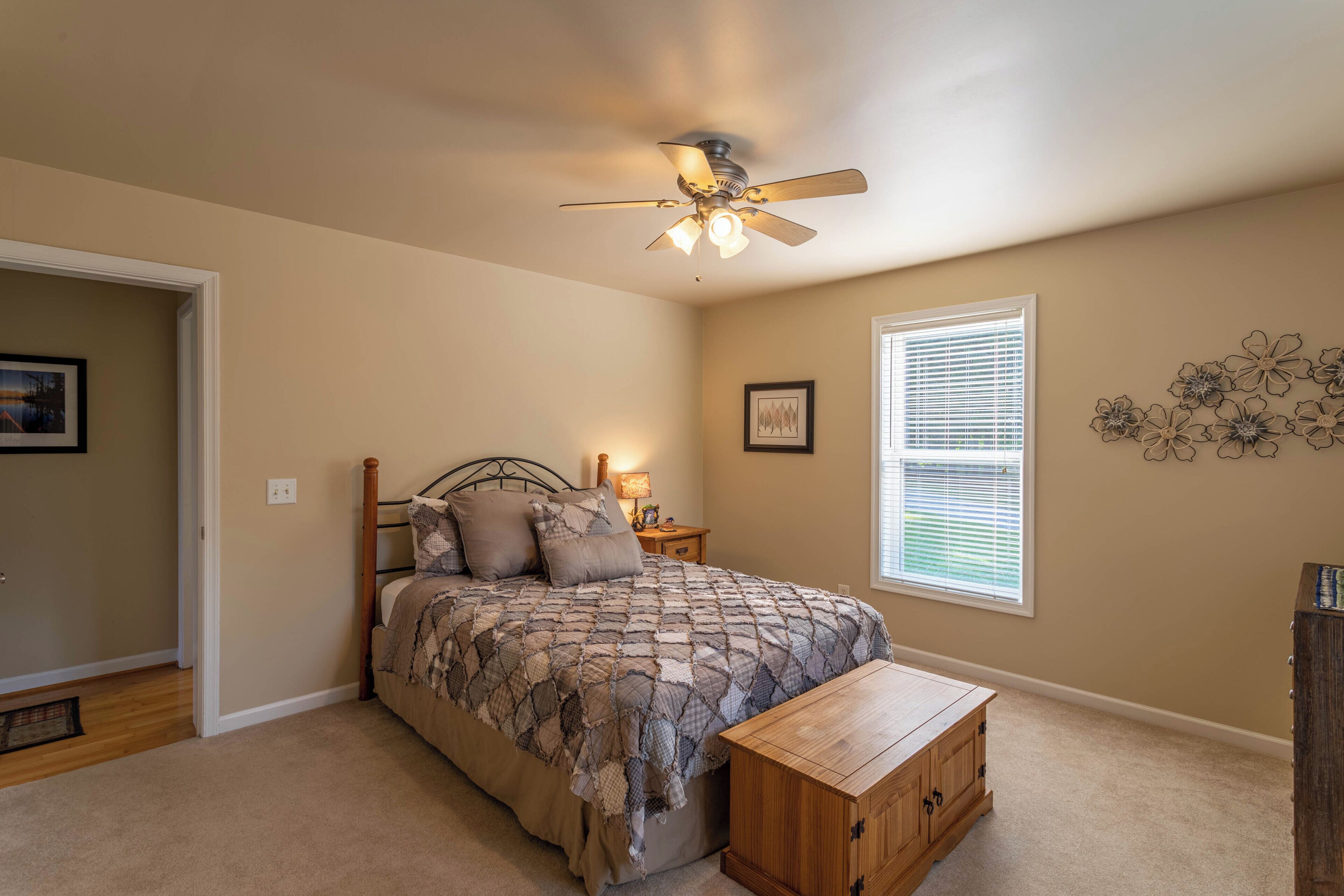A bedroom with a bed, a wooden nightstand, a ceiling fan, a window with blinds, and a decorative wall piece.