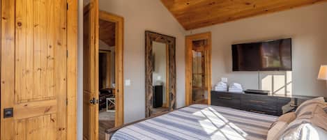 A bedroom with a wooden ceiling, a large bed with a striped bedspread, a flat-screen TV, and a dresser with towels.
