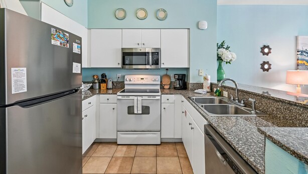 A modern kitchen with white cabinets, a stainless steel refrigerator, a microwave, an oven, and a granite countertop. - Shores of Panama 1323 (Panama City Beach)