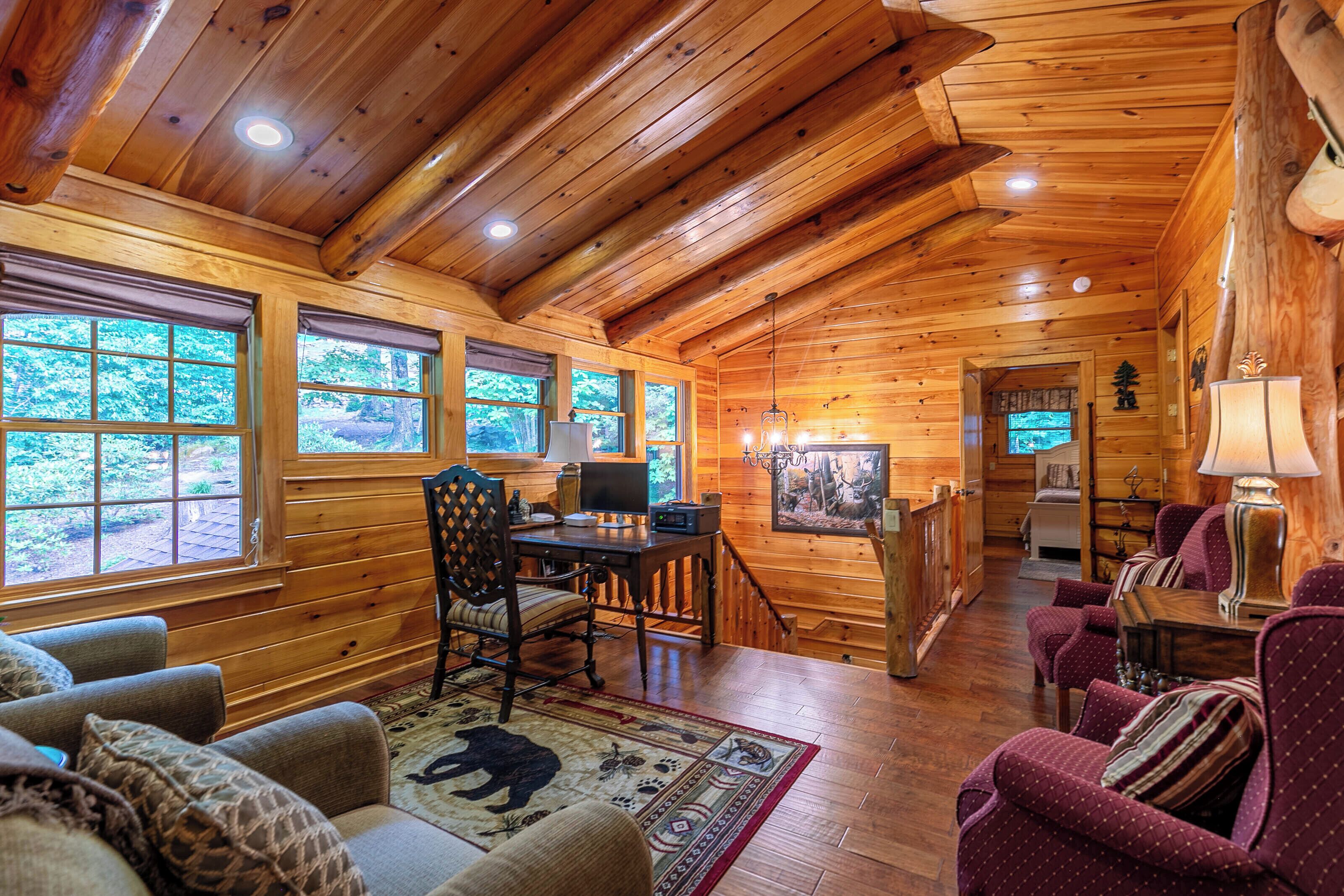 A log cabin interior with a wooden ceiling, large windows, a dining area, and a living room with sofas and a rug.