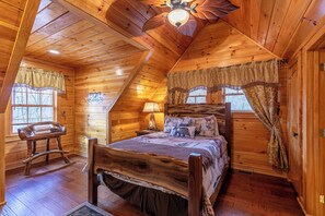 A cozy log cabin bedroom with a wooden bed, a nightstand, a lamp, and a window with curtains. - 4BR Running Waters Lodge at Eagles Nest (Banner Elk)
