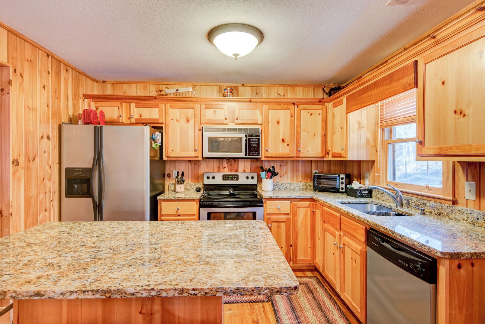 A kitchen with wooden cabinets, granite countertops, a refrigerator, a microwave, a stove, a dishwasher, and a window with blinds.
