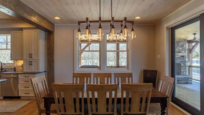 Ein Essbereich mit einem Holztisch und Stühlen, einer Chandelierleuchtre und großen Fenstern mit Blick ins Freie.