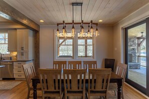 A dining area with a wooden table and chairs, a chandelier, and large windows with a view of the outdoors.