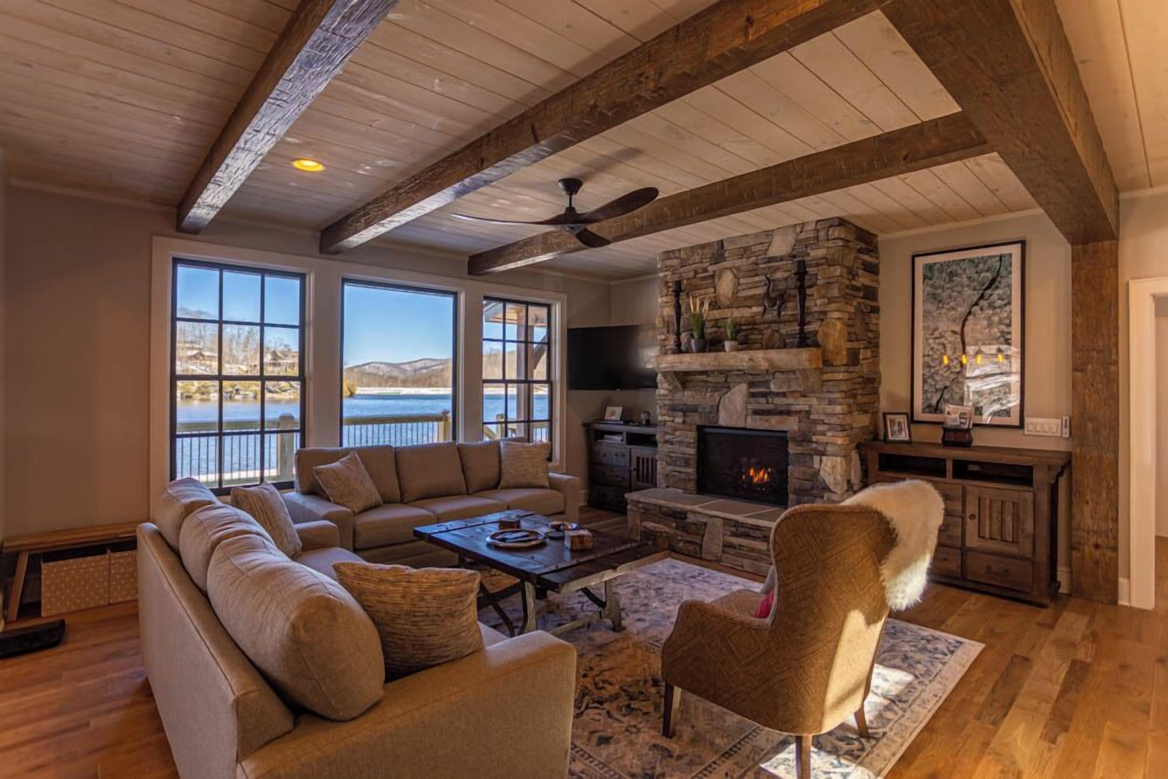 A spacious living room with a stone fireplace, large windows overlooking a body of water, and a cozy seating area.