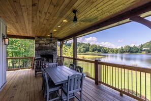 A covered deck with a stone fireplace, wooden furniture, and a view of a lake.