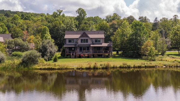 A large, two-story house with a wrap-around porch, situated next to a calm lake, surrounded by dense trees and a grassy area.