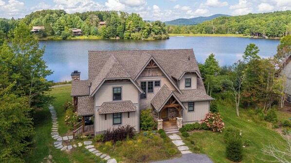 A large, two-story house with a steeply pitched roof, surrounded by a well-maintained garden and a lake.