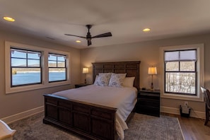 A bedroom with a large bed, two bedside tables, a ceiling fan, and two windows with views of a body of water.