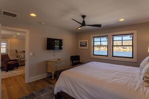 A bedroom with a large bed, a ceiling fan, two windows with a view, a dresser, and a TV on the wall.