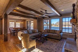 A spacious living room with a large sofa, a coffee table, and a fireplace. There are large windows offering a view of the outdoors.