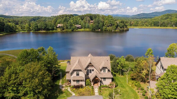 A large house with a stone foundation and a steeply pitched roof, surrounded by lush greenery and a lake.