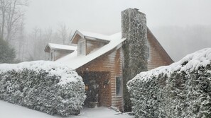 Una cabaña de troncos con techo nevado y chimenea, rodeada de arbustos cubiertos de nieve.