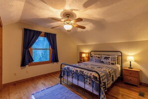 Un dormitorio con cama, ventilador de techo, ventana con cortinas, mesita de noche con lámpara y alfombra.