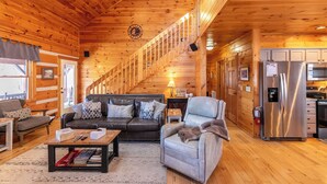 Een houten cabin interieur met een woonkamer, bank, fauteuil, salontafel, open haard en een keukenhoek.