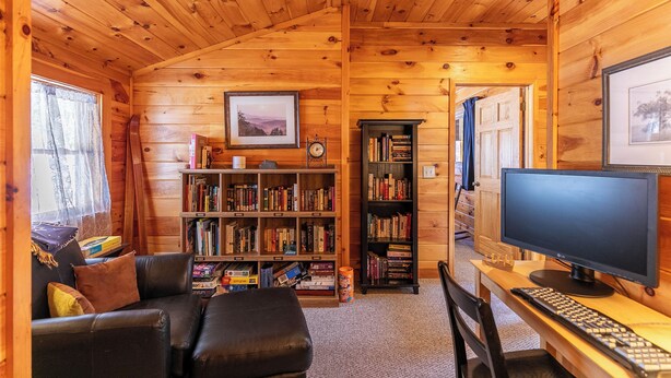 Een gezellig interieur van een blokhut, met een houten plafond, een boekenkast, een bureau met computer en een comfortabele zithoek.