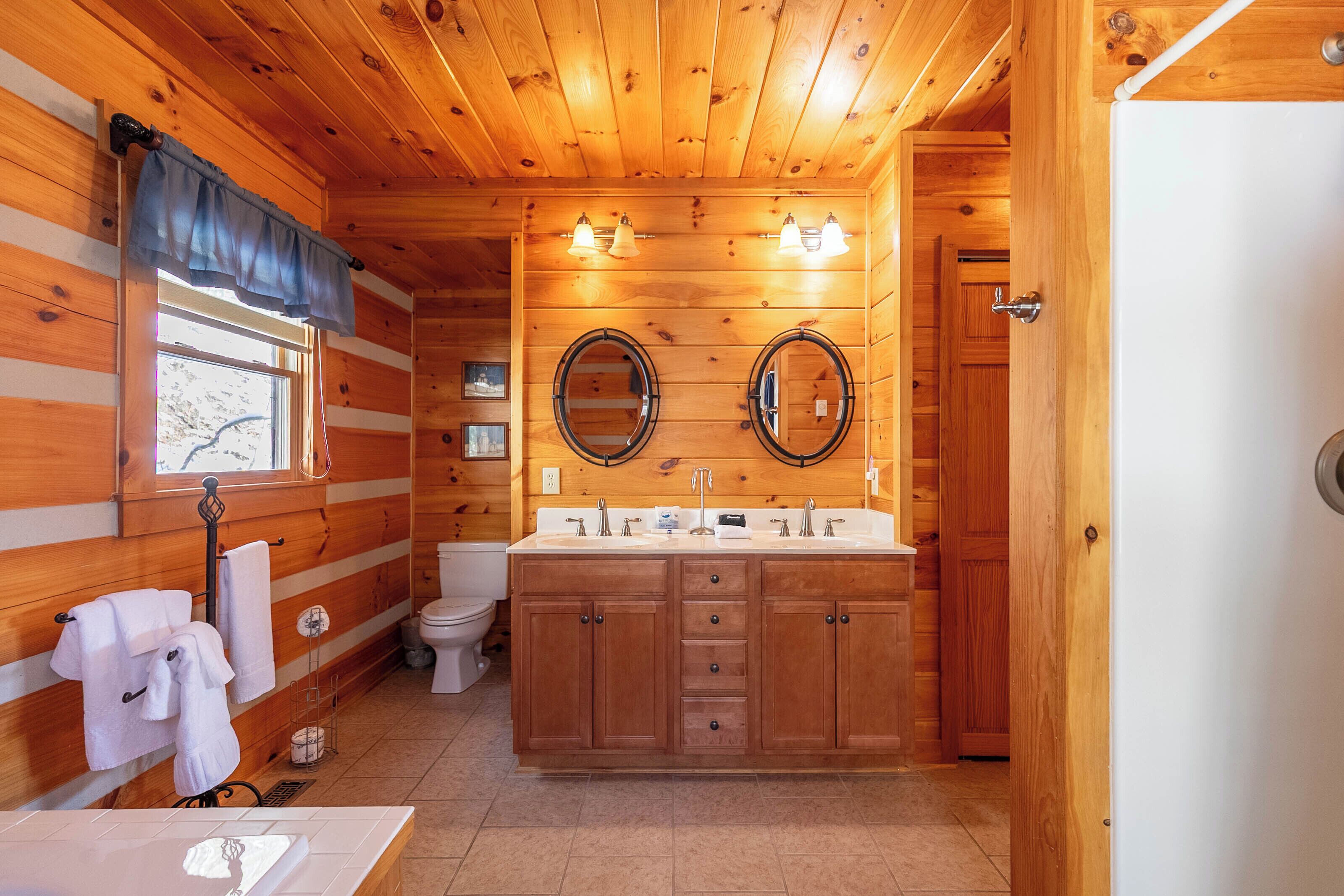 A wooden cabin bathroom with a double sink, two mirrors, a toilet, and a window with a curtain.