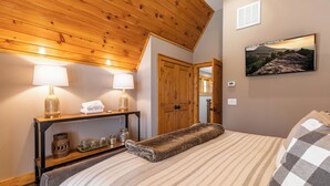 A bedroom with a wooden ceiling, a bed with striped bedding, two bedside lamps, a nightstand with decorative items, and a framed landscape painting on the wall. - Elk Bluff at Eagles Nest (Banner Elk)