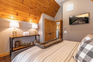 Ein Schlafzimmer mit Holzbalkendecke, einem Bett mit gestreifter Bettwäsche, zwei Nachttischlampen, einem Nachttisch mit Dekoration und einem gerahmten Landschaftsbild an der Wand.