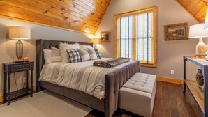 A bedroom with a wooden ceiling, a large bed with a striped blanket, two bedside tables with lamps, a window with blinds, and a painting on the wall. - Elk Bluff at Eagles Nest (Banner Elk)