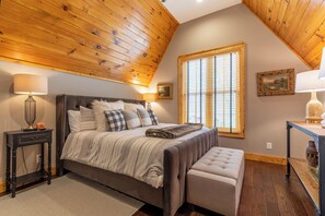 Ein Schlafzimmer mit Holzbalkendecke, einem großen Bett mit gestreifter Decke, zwei Nachttischen mit Lampen, einem Fenster mit Jalousien und einem Gemälde an der Wand.